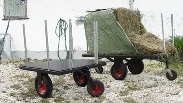 To store hø, står der to vogn med røde hjul og metalstænger på en grusbelagt gårdsplads.