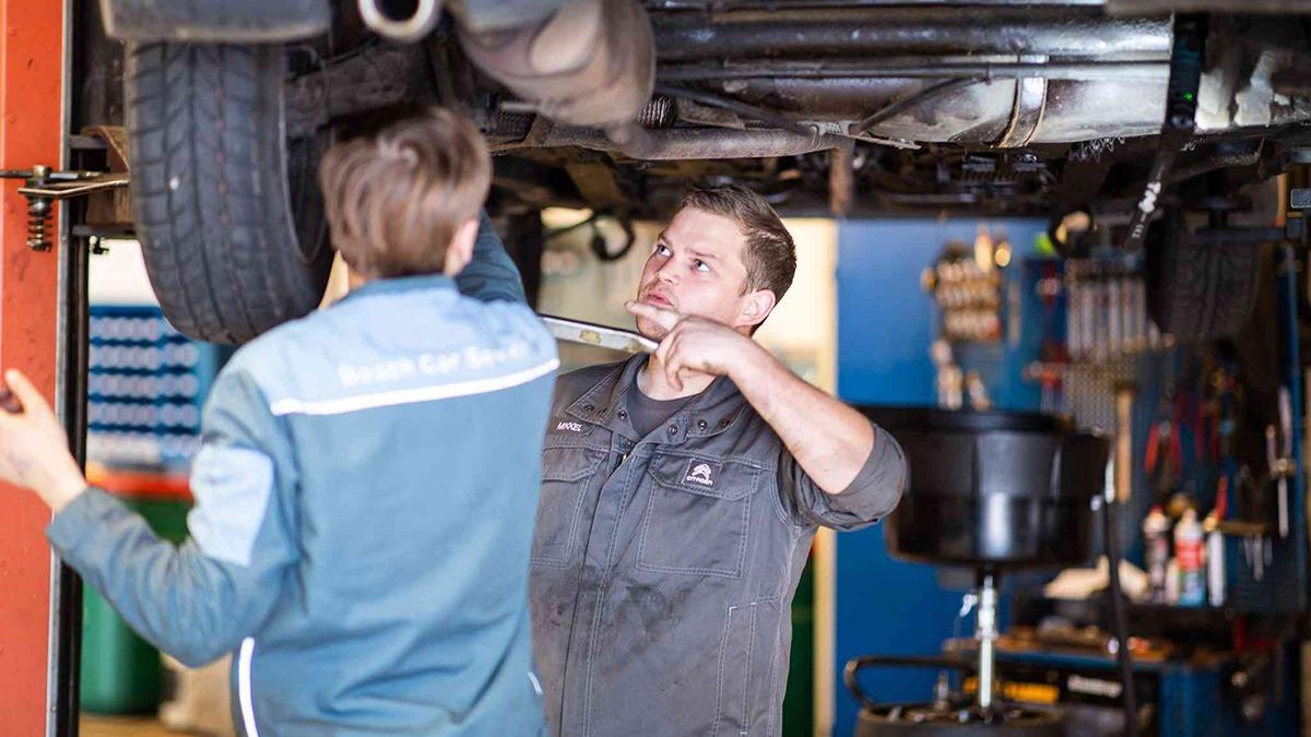 To mekanikere arbejder under en bil i et autoværksted, mens de udfører reparationer og vedligeholdelse.