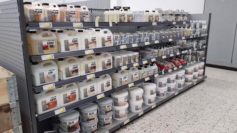 Hylder med forskellige malinger og træbeskyttelse i en butik, organiseret i klare beholdere og bøtter.