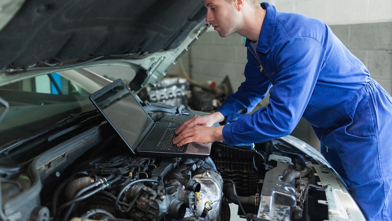 En mekaniker i blå overalls arbejder på en bilmotor med en bærbar computer.
