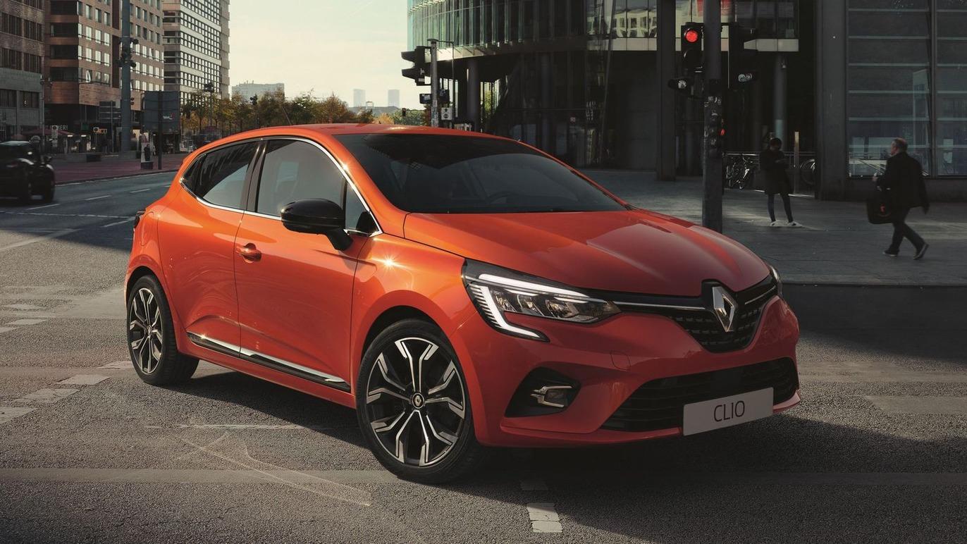 En rød Renault Clio parkeret ved et lyskryds i en by med moderne bygninger i baggrunden.