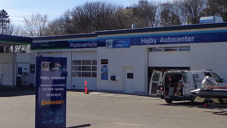 Højby Autocenter med værksted og serviceområde, beliggende i et industriområde med biler og skilt ved indgangen.