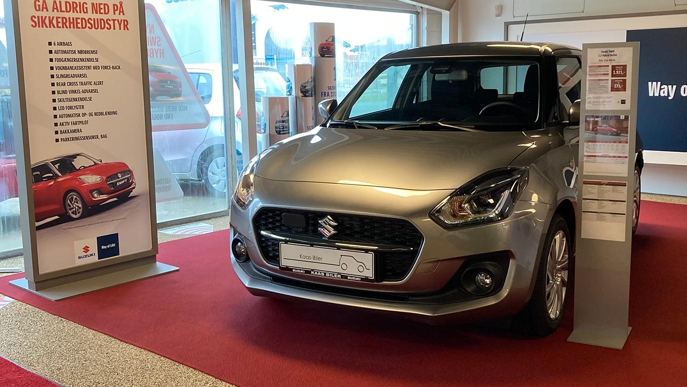 En sølvfarvet Suzuki bil udstillet i en bilforhandler med informationsplakater i baggrunden.