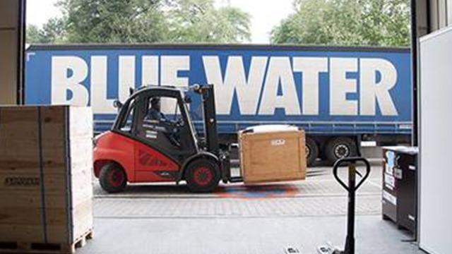 Gaffeltruck transporterer en kasse foran en stor lastbil med teksten "BLUE WATER" på siden.