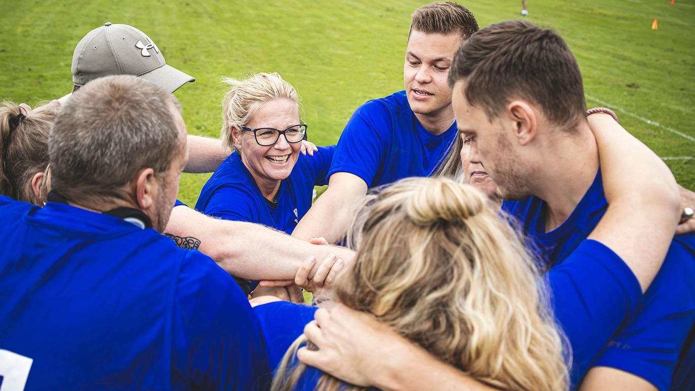 Gruppe af mennesker i blå t-shirts, der står i en cirkel og klapper hænderne sammen på en grøn græsplæne.
