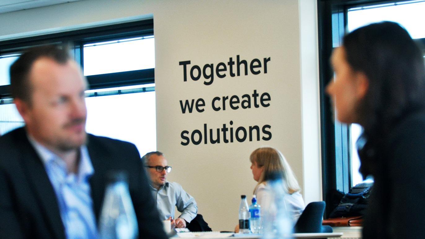 Møde i kontormiljø med fokus på samarbejde, hvor teksten "Together we create solutions" er synlig på væggen.