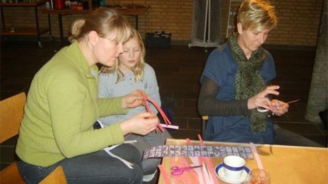 Tre personer sidder ved et bord og laver håndarbejde med farvet papir og saks.