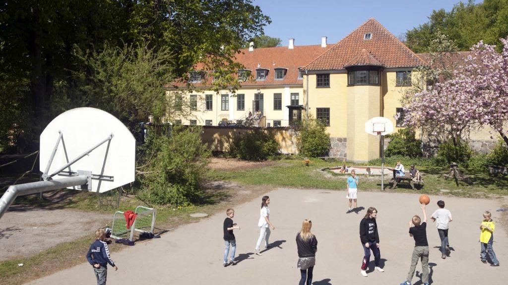 Børn spiller basketball på en legeplads foran en stor bygning med træer og blomster.