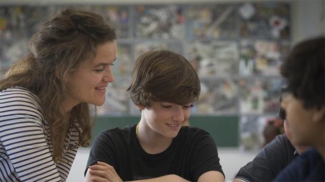 Lærer hjælper en dreng med opgaver i klasseværelset, mens en anden dreng lytter opmærksomt.
