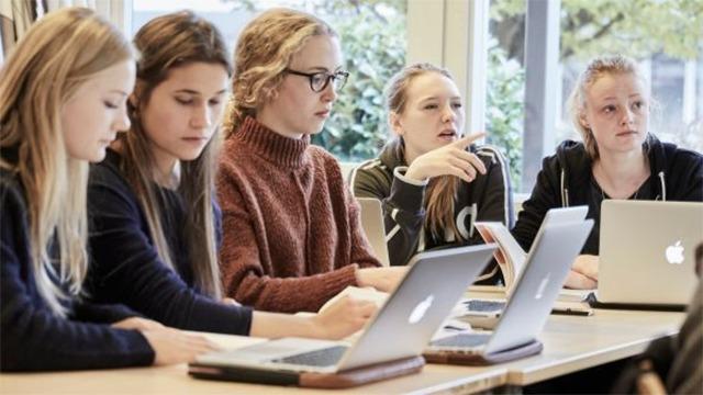 Fem unge kvinder sidder ved et bord med bærbare computere og diskuterer i et lyst klasseværelse.