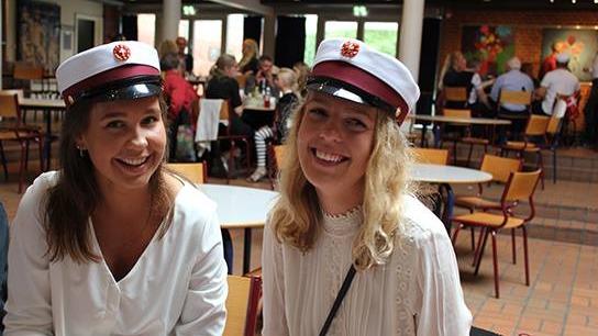 To unge kvinder iført studenterhuer smiler og sidder ved et bord i en festlig atmosfære.