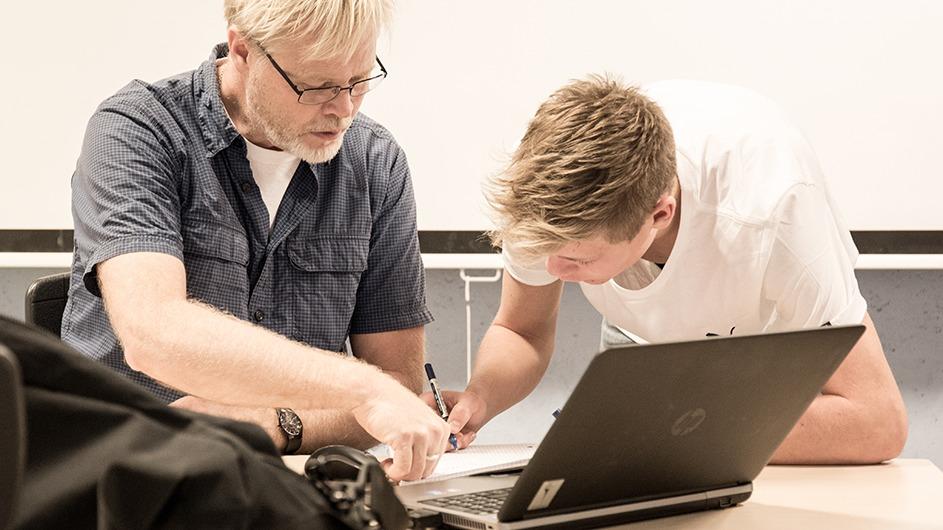 To mænd arbejder sammen ved en bærbar computer, hvor den ældre mand viser den yngre mand noget på papiret.