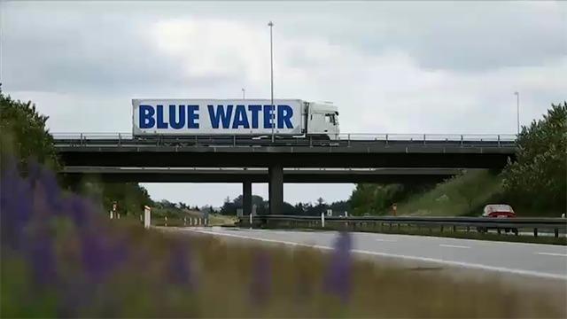 Lastbil med "BLUE WATER" på siden kører over en motorvejsbro med grønne marker i forgrunden.
