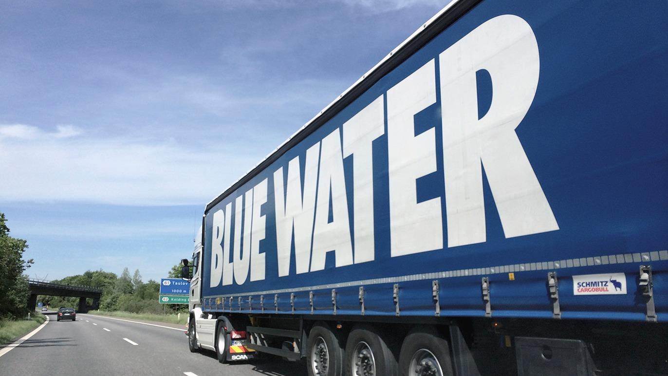 Lastbil med stor blå "BLUE WATER" tekst kører på motorvejen under en klar himmel.