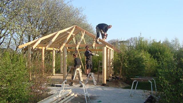 Tre håndværkere bygger et træhus med tagkonstruktion under en blå himmel og grønne træer i baggrunden.