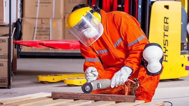 En arbejdstager i orange arbejdstøj og sikkerhedsudstyr, der bruger en slibemaskine på et stykke træ i et lager.