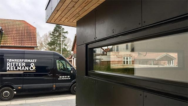 Billede af en sort varevogn med teksten "Tømrerfirmaet Ritter & Kelman" parkeret ved en moderne bygning.