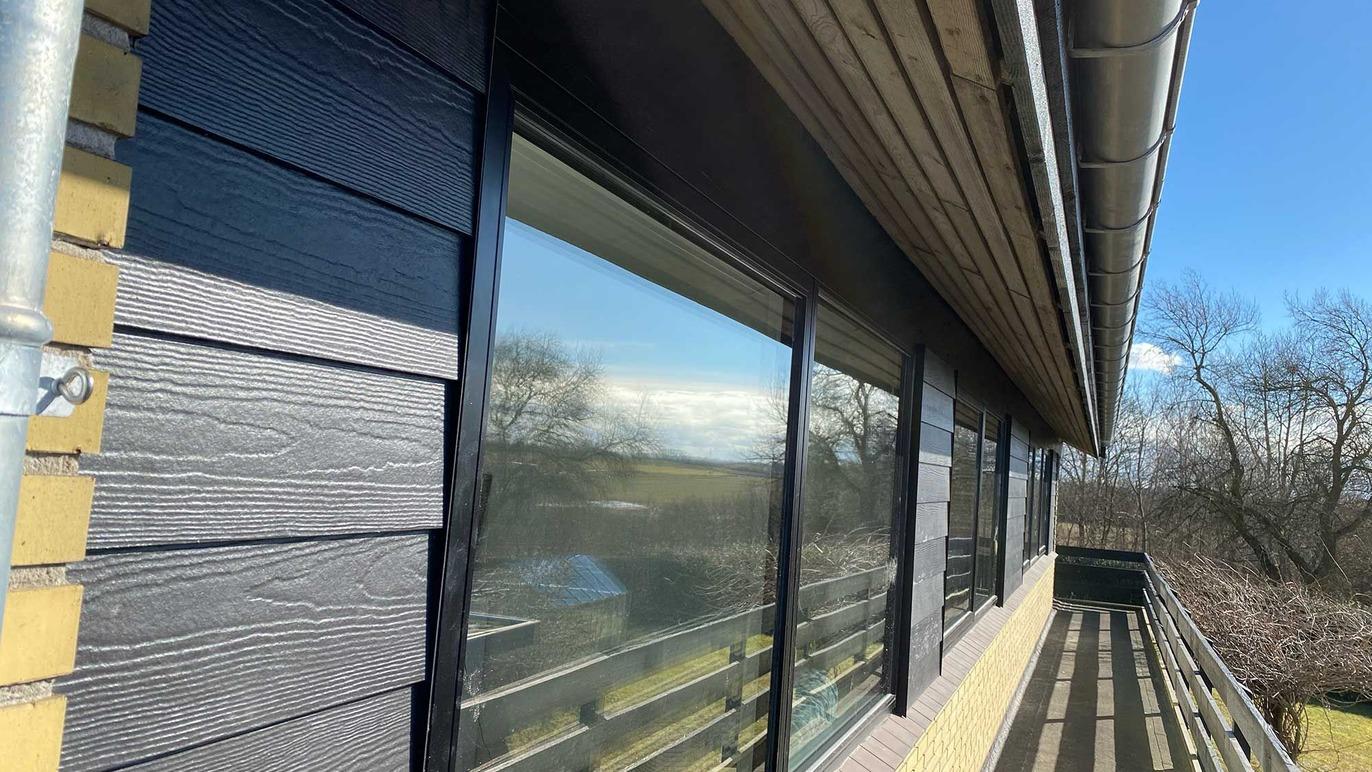 Husets facade med store vinduer og en balkon, omgivet af træer og en blå himmel.