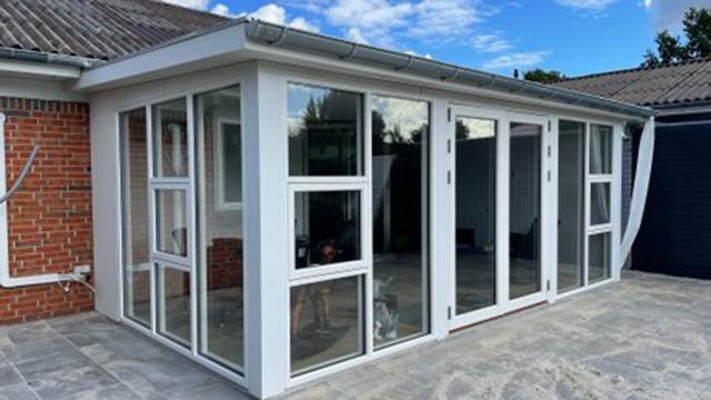 En moderne udestue med store vinduer og glasdøre, bygget i hvidt træ og placeret på en flisebelagt terrasse.