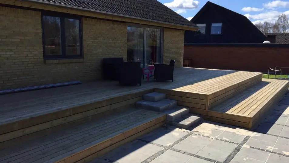 Hus med træterrasse og trapper, omgivet af fliser og en klar blå himmel.