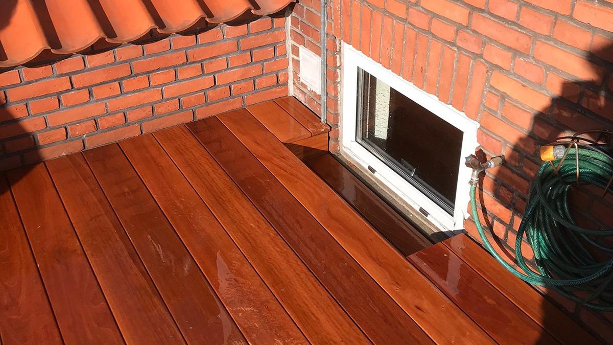 Nybehandlet terrasse med glat træbelægning og murstensvæg, der har et vindue og en vandslange.