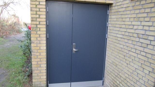 En mørkeblå dør med metalhåndtag på en murstensvæg i en have.
