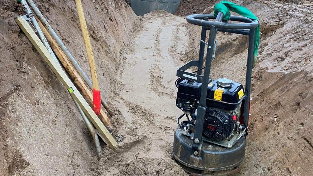En komprimeringsmaskine står i en udgravning med værktøj som spade og planker ved siden af.