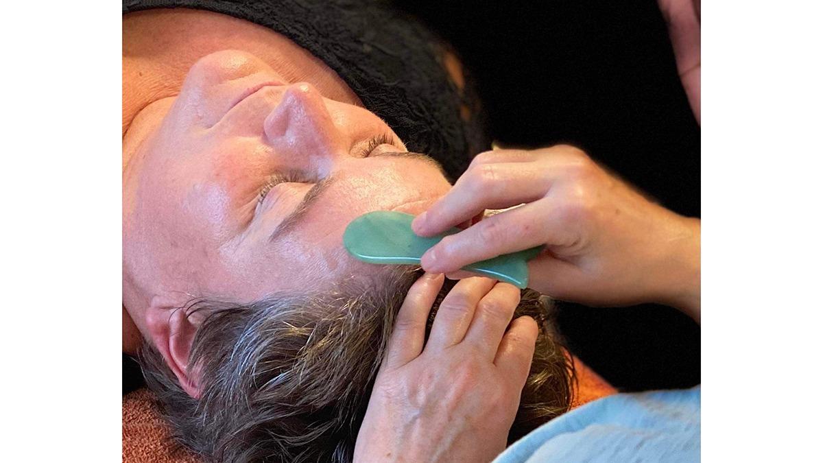 Kvinde får ansigtsbehandling med guasha værktøj, mens hun ligger afslappet på en briks.