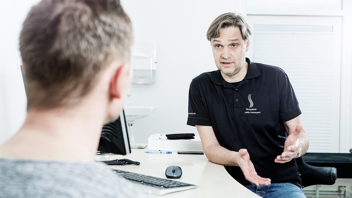 En læge taler med en patient i et moderne kontor med computer og medicinsk udstyr.