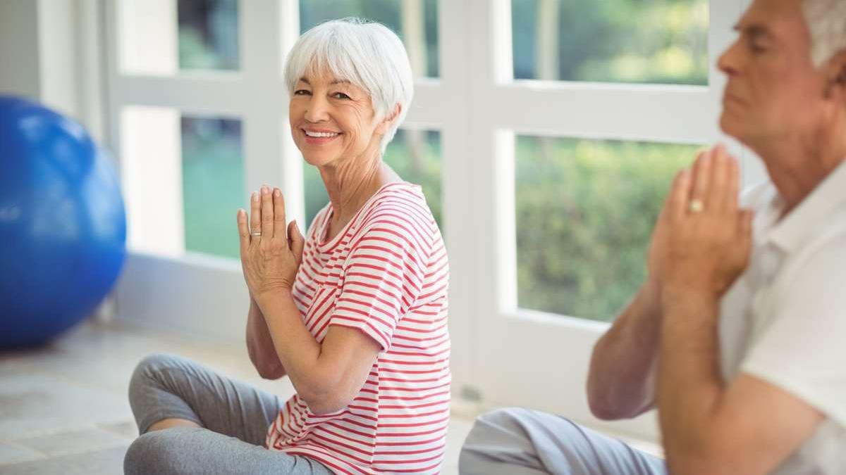 Ældre par, der praktiserer yoga indendørs, smilende med hænderne i bøn, i et lyst rum med en stor blå bold.