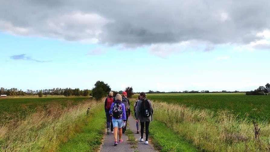 Gruppe af vandrere på en grusvej omgivet af marker og skyer i baggrunden.