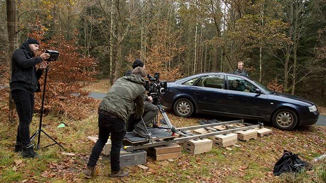 Filmoptagelse i skoven med en bil, kameraudstyr og personer, der arbejder på en scene.