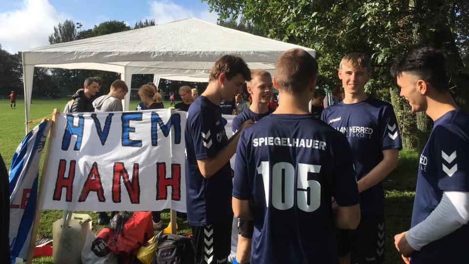 Gruppe unge fodboldspillere står under et telt med en banner, der siger "HVEM HAN H".