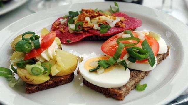 En tallerken med smørrebrød, kartofler, æg, tomater og en rød pølse med løg og grønne urter.