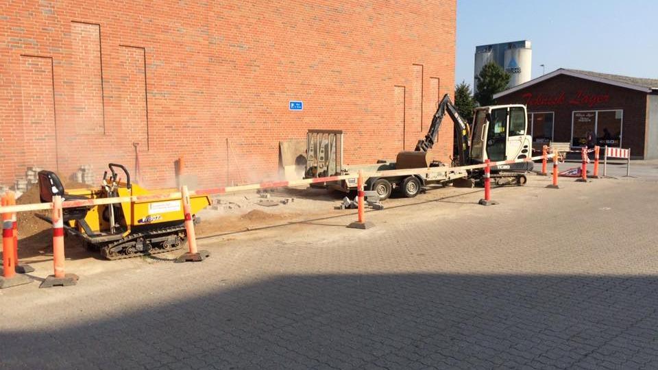 Byggeplads med gravemaskine og asfaltmaskine ved en murstensvæg, omgivet af afspærringsbånd og advarselsskilt.