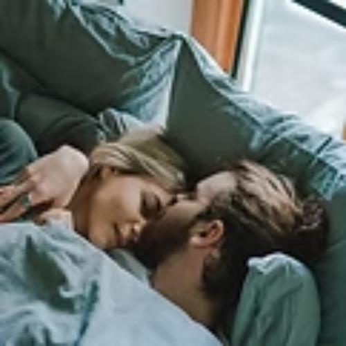 Et par ligger tæt sammen i sengen, omfavnet under bløde dyner, mens de deler et øjeblik af intimitet.