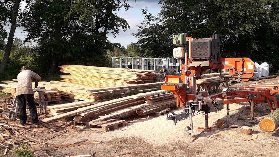 En mand arbejder med en træbearbejdningsmaskine ved en stak af tømmerplanker i et skovområde.