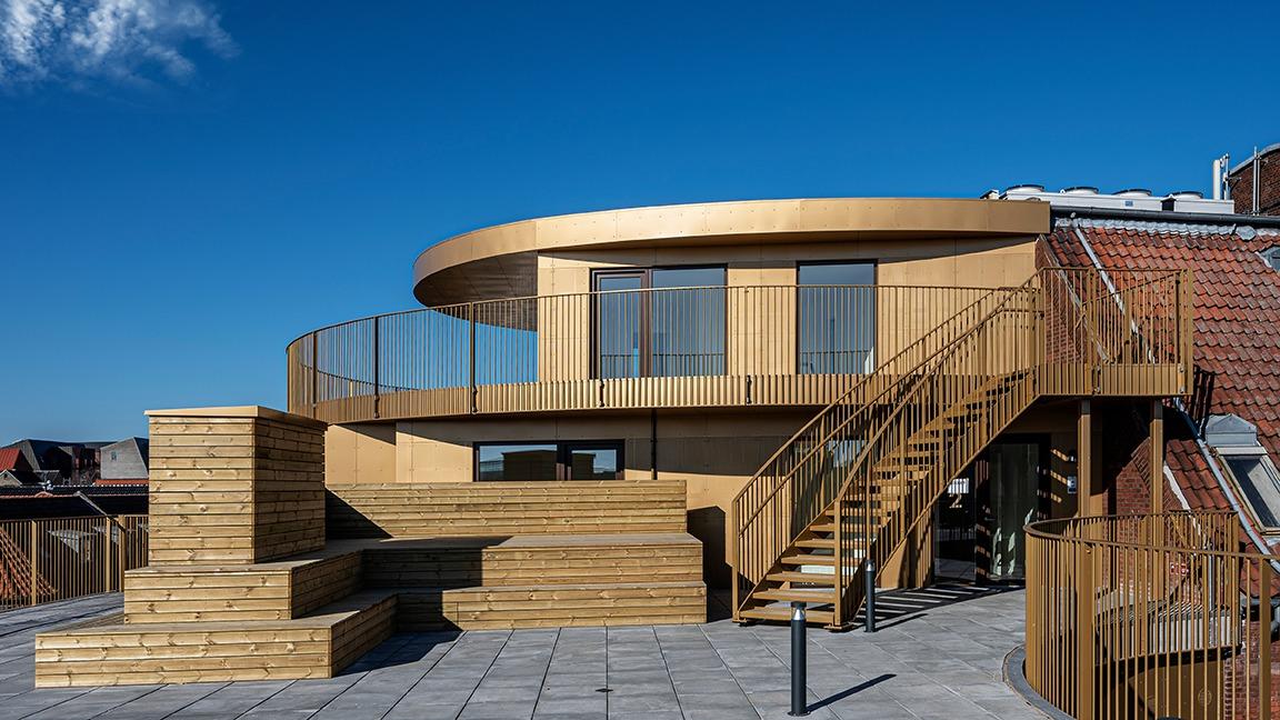 Moderne træhus med rund terrasse og trappe, beliggende på en tagterrasse under en klar blå himmel.