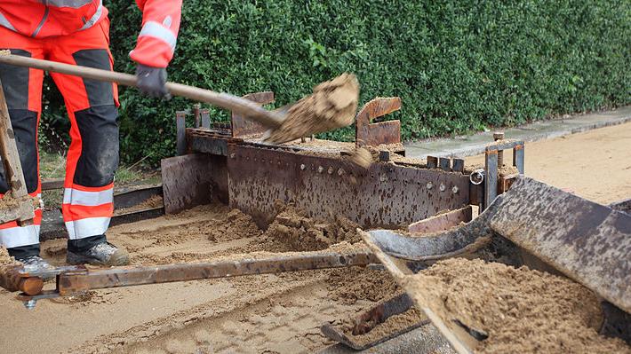 En arbejdstager i refleksjakke graver sand op med en skovl ved en byggeplads.