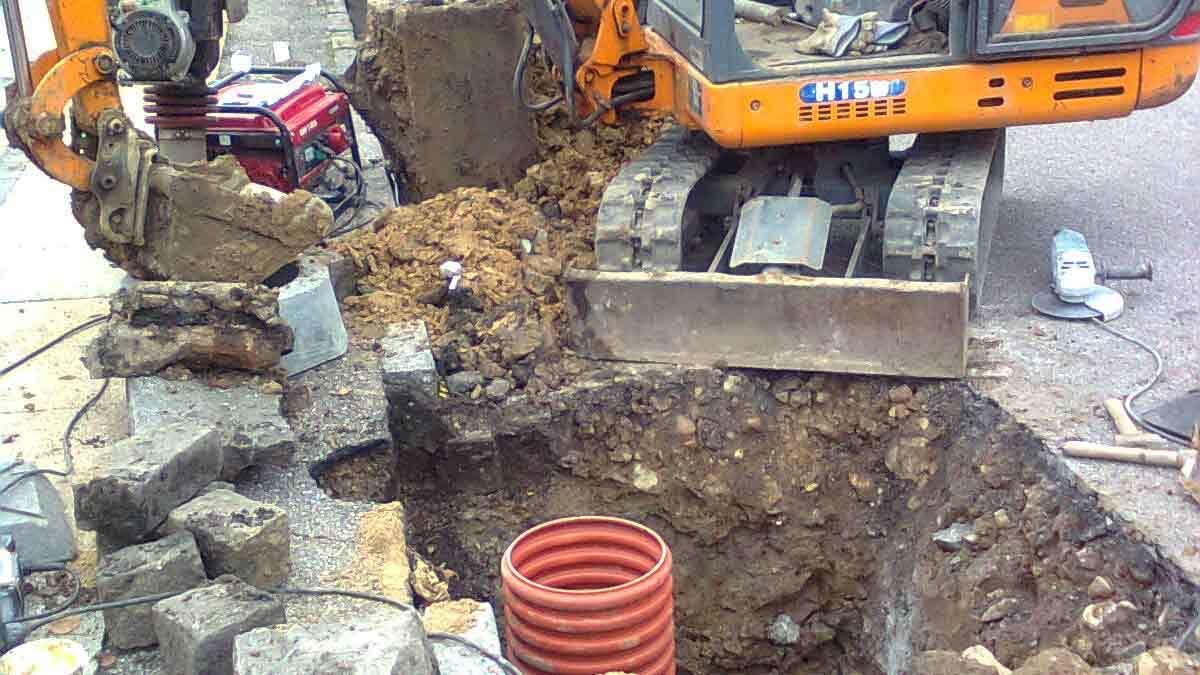 En gravemaskine arbejder på en byggeplads med en stor gravet grøft og synlige rør og sten.