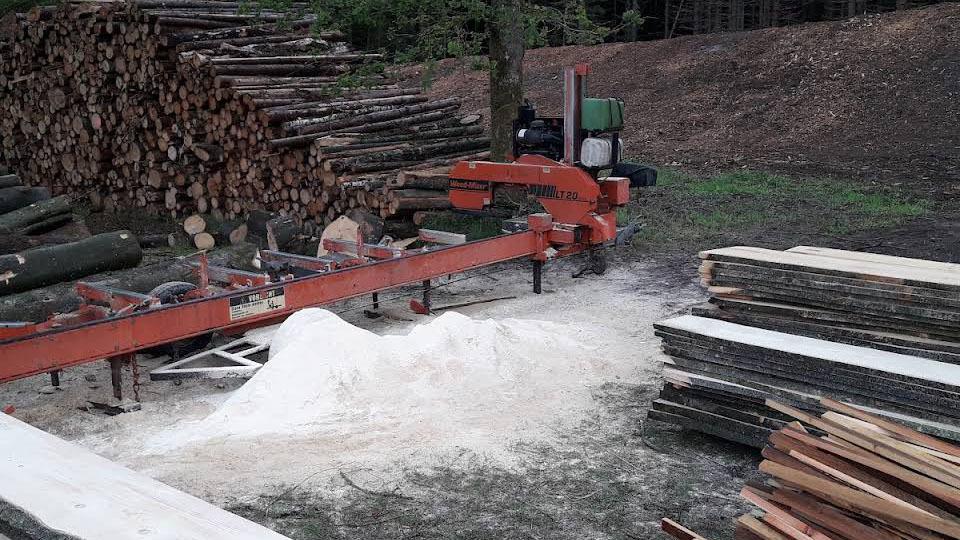 En træbearbejdningsmaskine står ved siden af stakke af tømmer og savsmuld på en skovgrund.