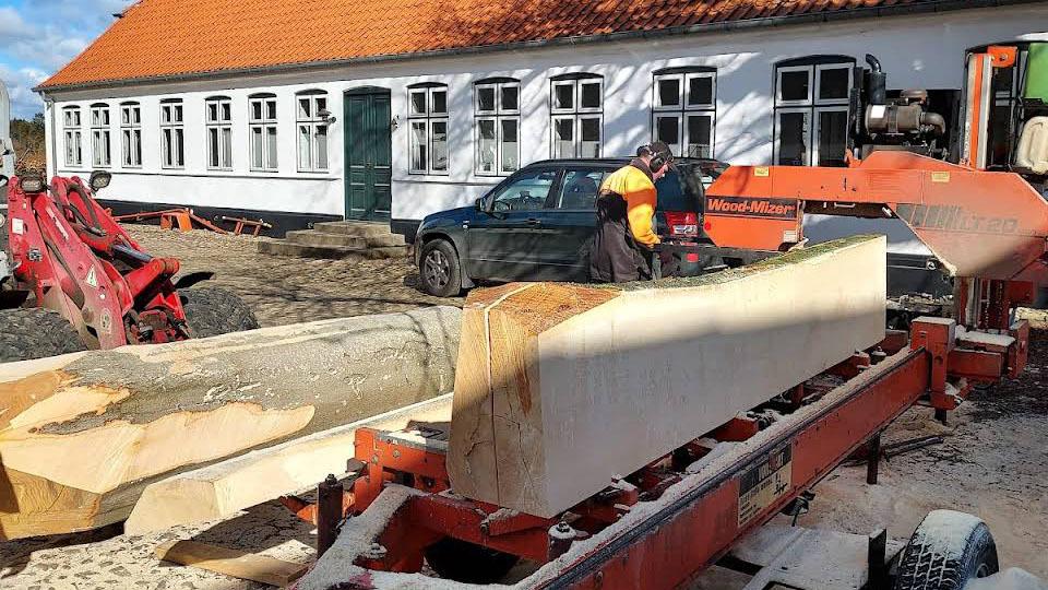 En mand arbejder med en træskærer ved en bygning, mens en bil holder i baggrunden.