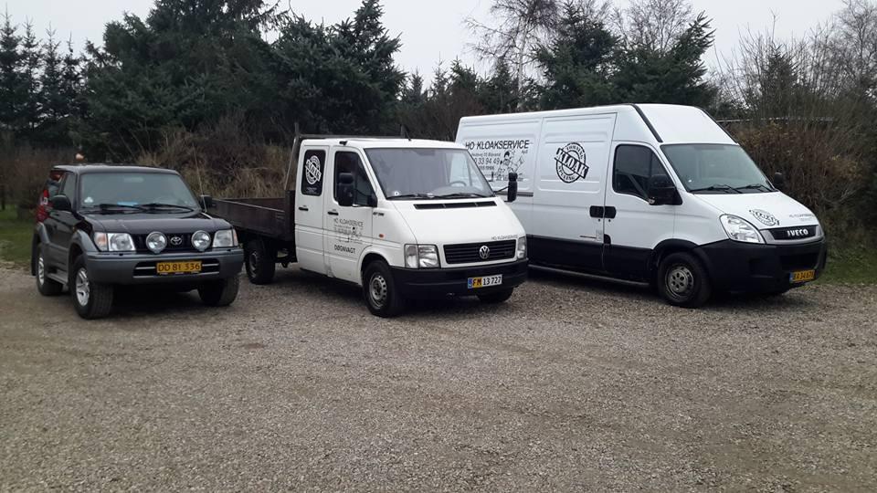 Tre biler parkeret på en grusvej, herunder en sort SUV, en hvid varevogn og en hvid pickup.