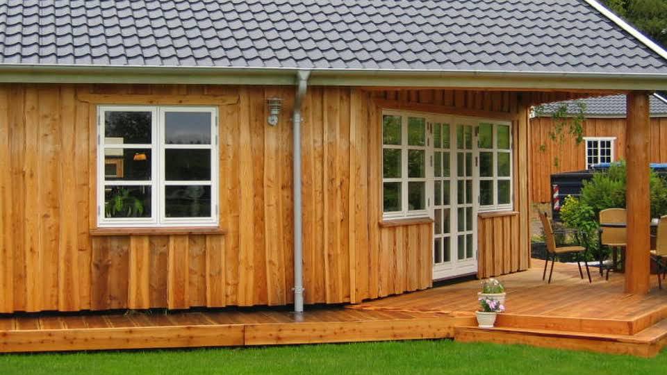 Et træhus med en terrasse, vinduer og en dør, omgivet af grøn græsplæne.