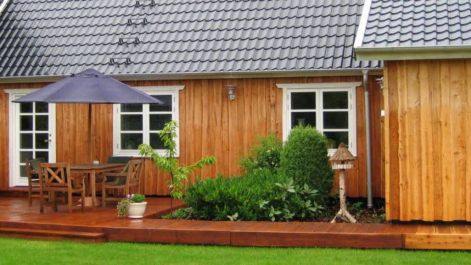 Et hyggeligt hus med træfacade, terrasse, havemøbler og en parasol i haven.