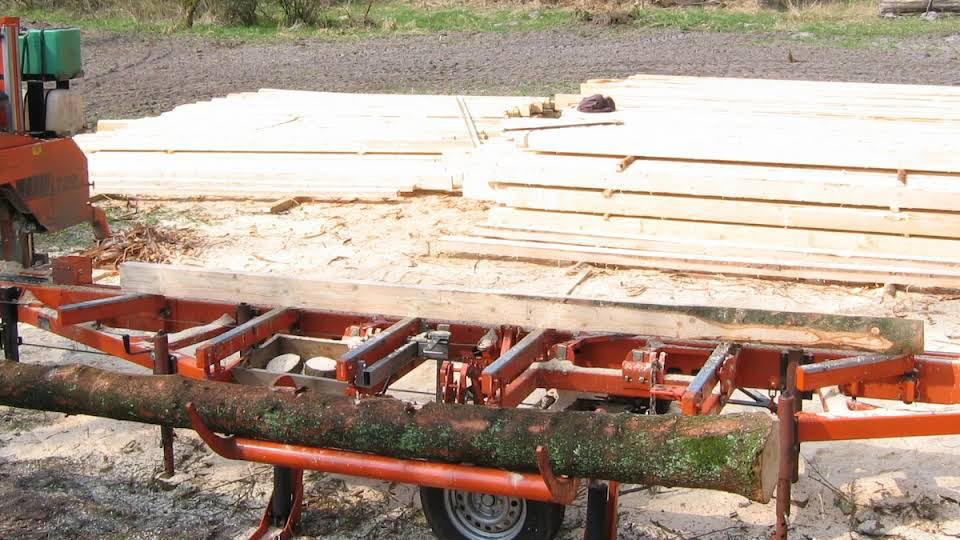 En træbearbejdningsmaskine med en stamme og planker i baggrunden på en skovbrugsplads.