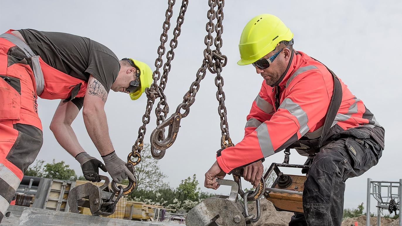To arbejdere i sikkerhedstøj og hjelme, der arbejder med kæder og løfteudstyr på en byggeplads.
