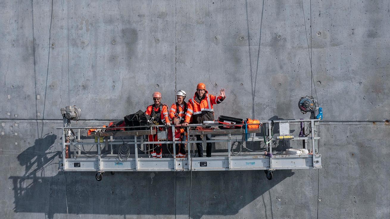 Tre arbejdere i orange sikkerhedstøj står på en stilladsplatform ved en betonvæg.