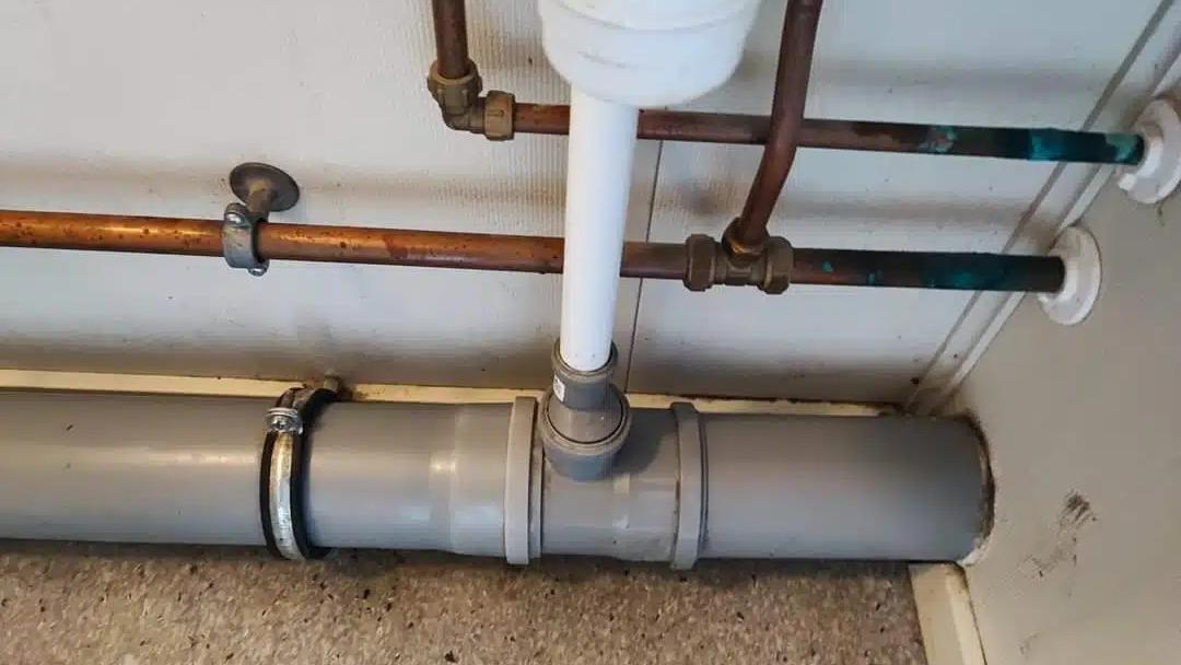 Rørinstallation med kobber- og plastledninger samt en grå afløbsrør i et værelse.
