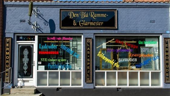 Butikken "Den Blå Ramme- & Glarmester" med farverige skilte og vinduer, der viser forskellige glasprodukter og tjenester.